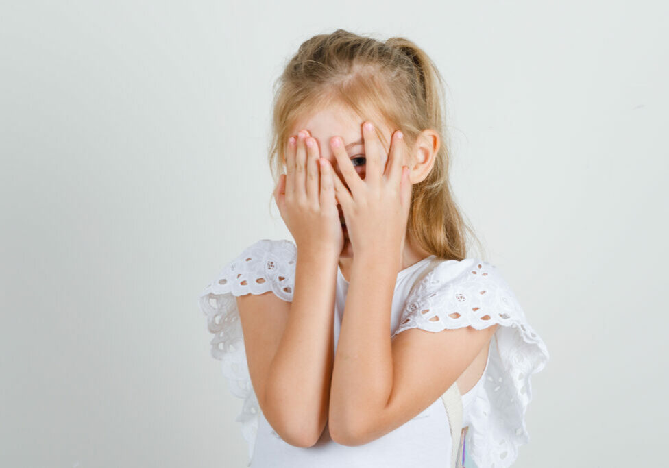 Little girl in white t-shirt, skirt looking through fingers with one eye , front view.